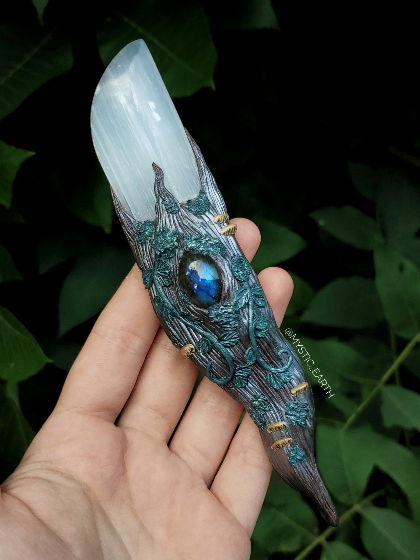 A hand sculpted athame dagger. The blade is non functional, made from a selenite crystal (glowing white stone). The handle is done in a woodland fairy style, of a winding tree branch that wraps around the sides of the blade and comes to a swirling point. There is a labradorite crystal centered just below the blade, surrounded by vines, moss, and realistic mushrooms.