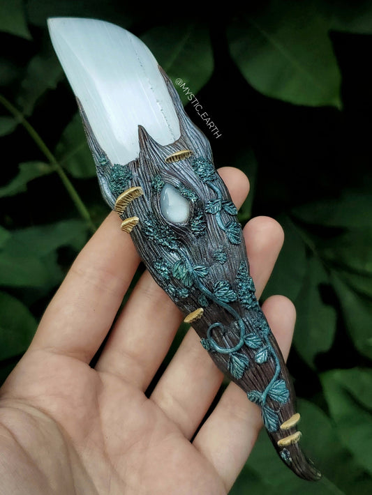 A hand sculpted athame dagger. The blade is non functional, made from a selenite crystal (glowing white stone). The handle is done in a woodland fairy style, of a winding tree branch that wraps around the sides of the blade and comes to a swirling point. There is a white moonstone crystal centered just below the blade, surrounded by vines, moss, and realistic mushrooms.