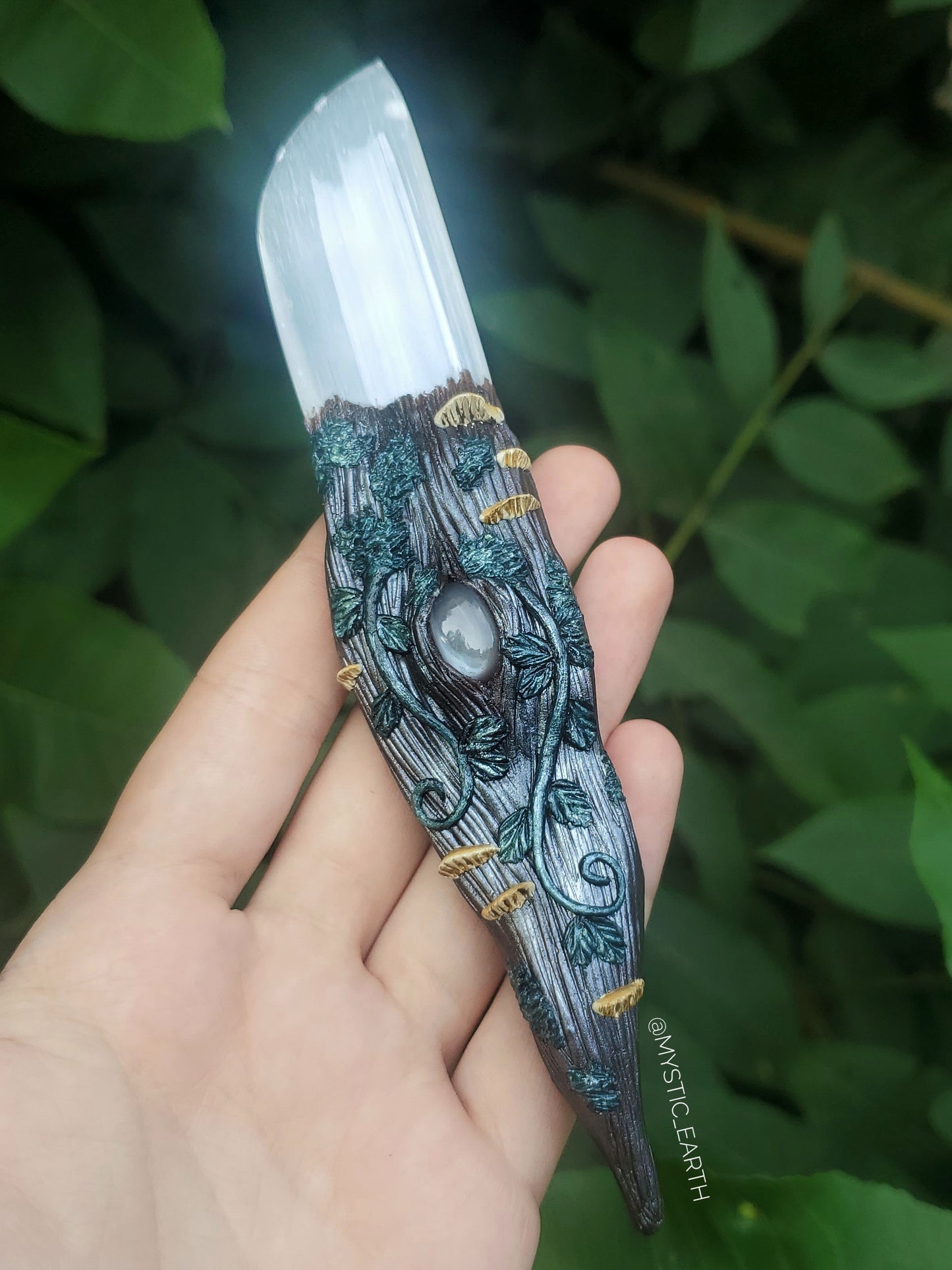 A hand sculpted athame dagger. The blade is non functional, made from a selenite crystal (glowing white stone). The handle is done in a woodland fairy style, of a winding tree branch that wraps around the sides of the blade and comes to a swirling point. There is a gray moonstone crystal centered just below the blade, surrounded by vines, moss, and realistic mushrooms.