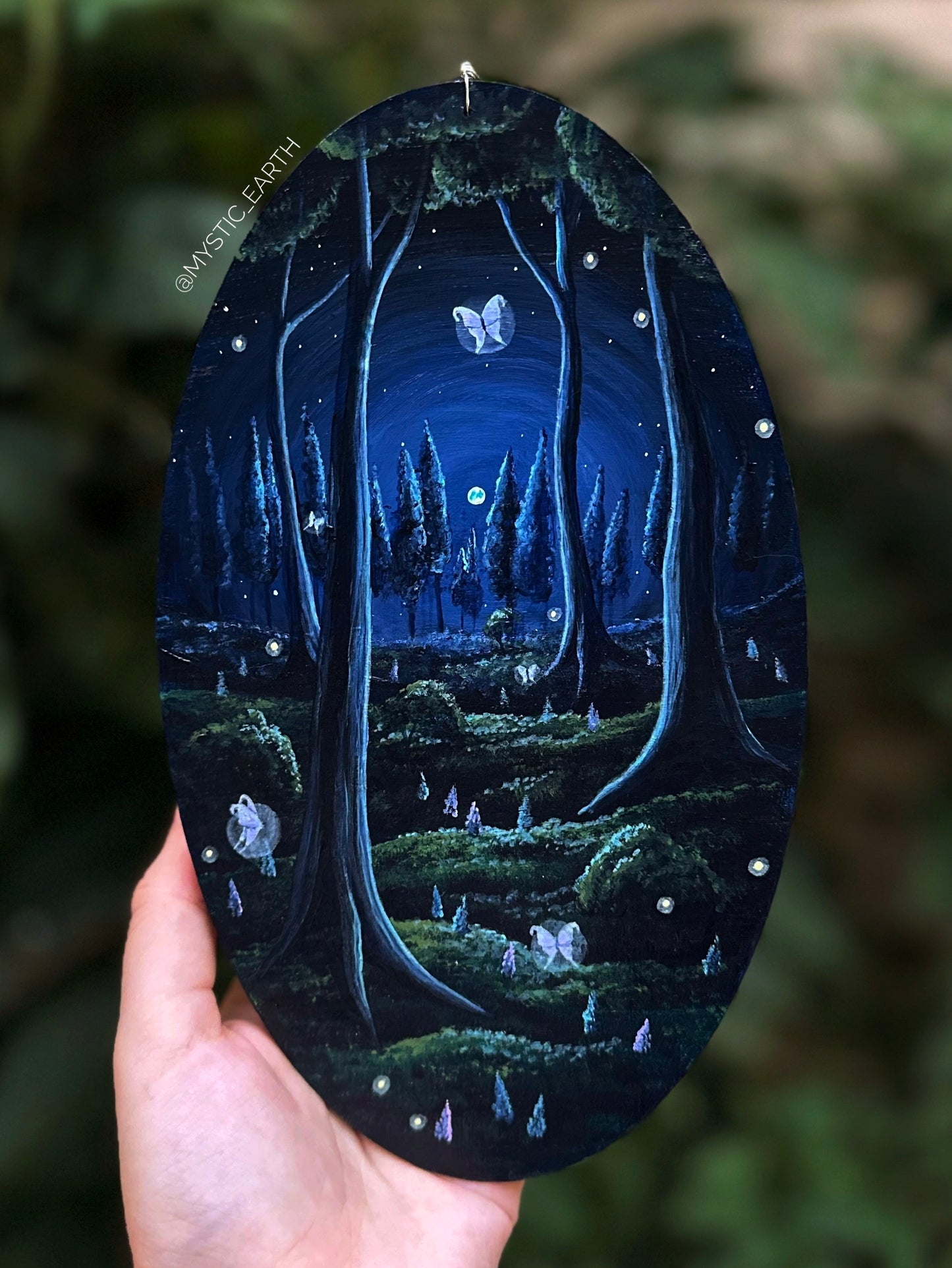 Oval-shaped acrylic painting on wood that hangs by a silver chain and loop. The painting depicts a dark, gloomy forest in blue and green shades. The sky is filled with stars and a full moon, with trees shown far away in the distance. Up close you can see the trunks of four trees. The grass is filled with hyacinth flowers, bushes, fairies, and fireflies.