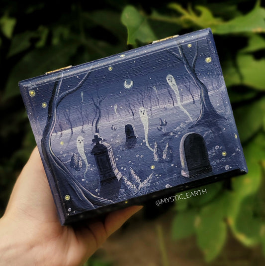 Wooden rectangle shaped box painted in a muted purple depicting a cemetery in a forest. There are five ghosts floating in the forest with fireflies. The tree are bare and cascade into the distance