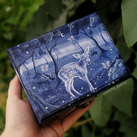 A deep, dark blue wooden box painted on top to depict a baby deer in a forest. It stands in the front center surrounded by fireflies and fairies. It is a nightime scene, the moon and stars are out