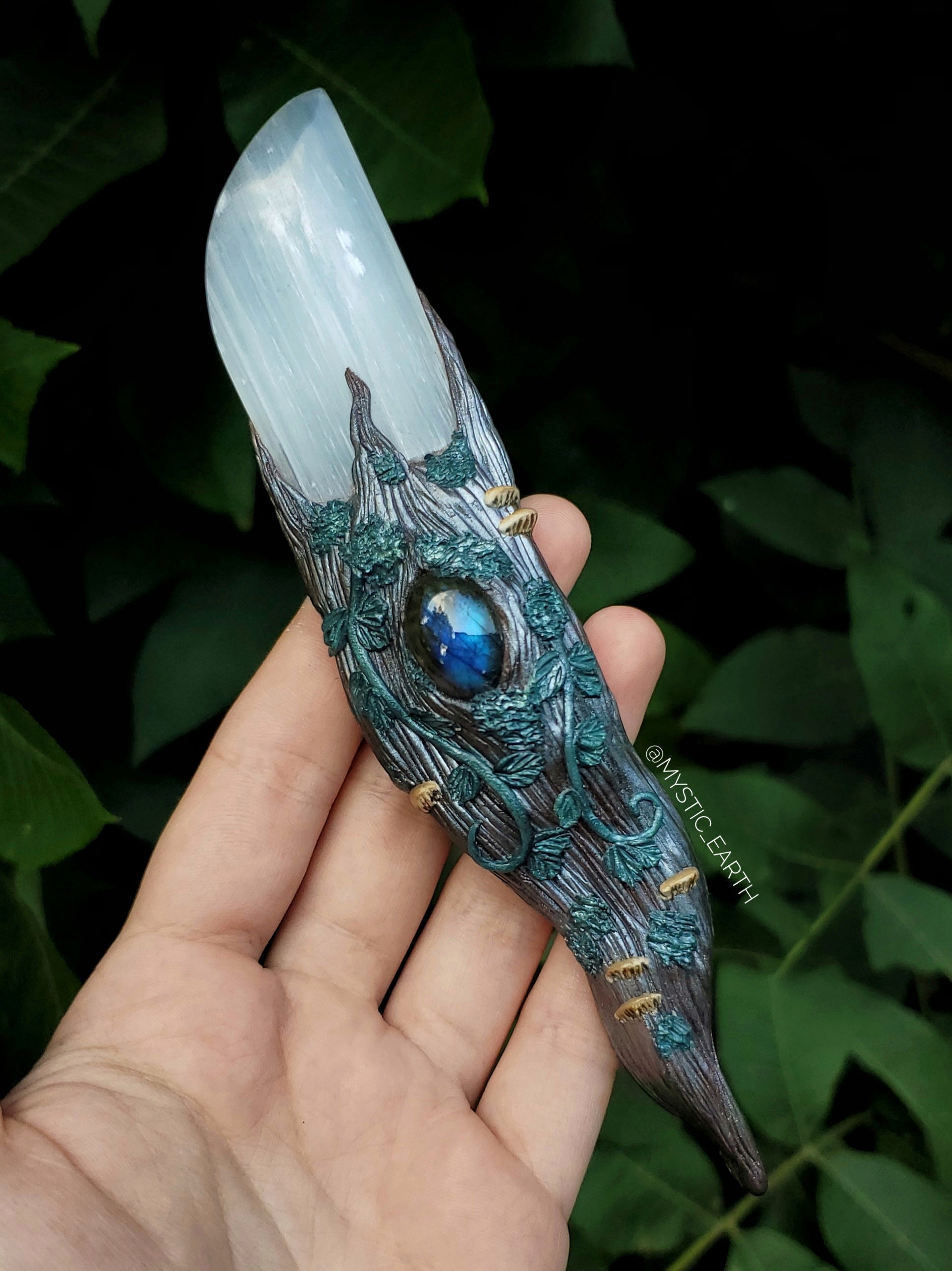 A hand sculpted athame dagger. The blade is non functional, made from a selenite crystal (glowing white stone). The handle is done in a woodland fairy style, of a winding tree branch that wraps around the sides of the blade and comes to a swirling point. There is a labradorite crystal centered just below the blade, surrounded by vines, moss, and realistic mushrooms. 