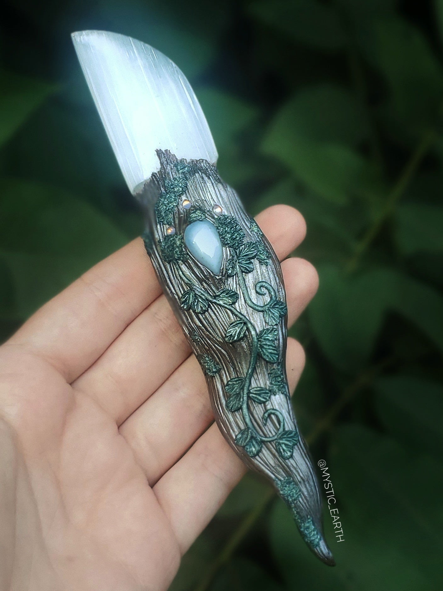 A hand sculpted athame dagger. The blade is non functional, made from a selenite crystal (glowing white stone). The handle is done in a woodland fairy style, of a winding tree branch that wraps around the sides of the blade and comes to a swirling point. There is a blue opal crystal centered just below the blade, surrounded by vines, moss, and iridescent glass gems. 